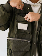 詳細画像
「胸元のロゴ刺繍とアシンメトリーな多機能ポケットのディテール特写。YKKカスタムジッパーとロゴ入りスナップボタンが、高品質のディテールを引き立て、機能性とスタイルを両立させています。ダブルレイヤーのリブ編みが施され、袖口と裾のフィット感と耐久性を向上させています。」
モデル画像
「モデルがアシンメトリーデザインの多ポケットキャンプワークジャケットを着用し、アウトドアシーンでのコーディネートを披露。ショート丈のルーズフィットシルエットが、カジュアルな日常コーディネートやアウトドア活動に最適なスタイルを提供します。」