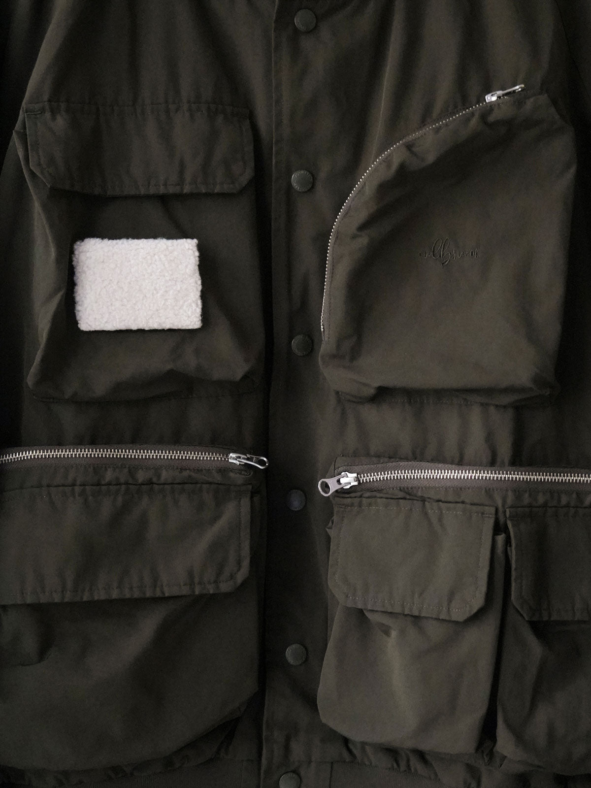 製品画像
「アシンメトリーデザインの多ポケットキャンプワークジャケット全体の展示図。クラシックなフィッシングジャケットとワークスタイルが融合したデザインで、ユニークなアシンメトリーデザインが実用性と個性を強調。防撥水機能を備えたポリエステルとコットン混紡生地を使用し、快適な着心地を提供します。」
詳細画像
「胸元のロゴ刺繍とアシンメトリーな多機能ポケットのディテール特写。YKKカスタムジッパーとロゴ入りスナップボタンが、高品質のディテールを引き立て、機能性とスタイルを両立させています。ダブルレイヤーのリブ編みが施され、袖口と裾のフィット感と耐久性を向上させています。」