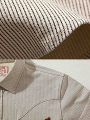 製品画像: アイボリー色のスナップボタン付きストライプシャツの正面図。ヴィンテージウエスタンデザインが特徴。
詳細画像: シャツの肩部ディテールとトライアングルレザー補強部分のクローズアップ。ダブルステッチと色織り生地が耐久性とスタイルを強調。