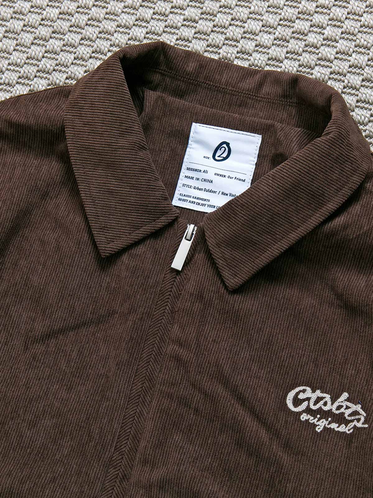 製品画像: レトロなコーデュロイジャケット平置き。シンプルな刺繍ロゴと襟付きデザインが特徴の秋冬必須アイテム
詳細画像: コーデュロイ素材の織り目と刺繍ロゴのディテール。保温性と軽量感を備えたジャケット