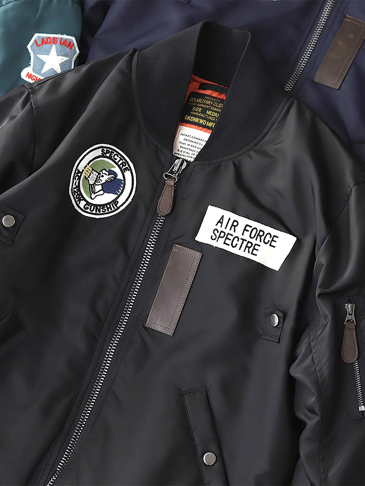 製品画像:ヴィンテージMA-1フライトジャケットの正面図。防水ナイロン素材とオレンジ色の裏地、刺繍バッジが特徴的なデザイン。
詳細画像:刺繍バッジと識別テープ、ジッパーのディテールをクローズアップした画像。ミリタリーウェアの本格的なスタイルを強調。