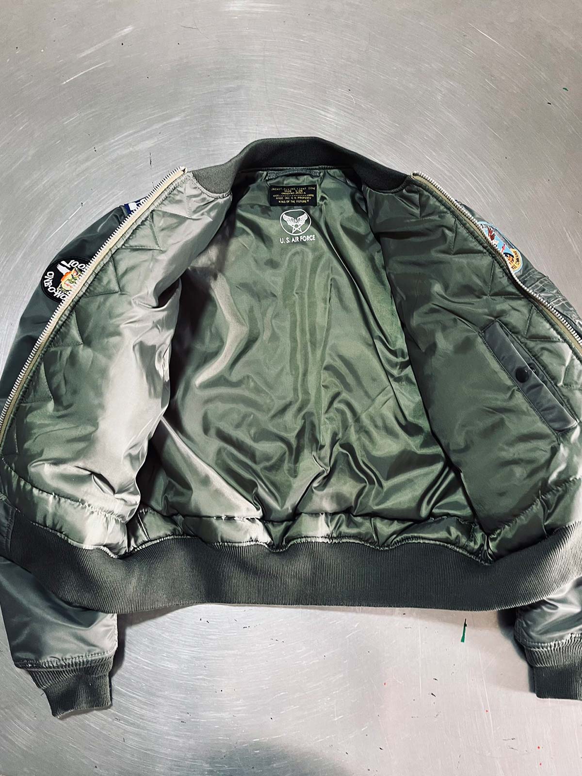 製品画像: L2-Bヴィンテージフライトジャケットの全体画像、ミリタリーグリーンとキャプテンランクの肩章が特徴のデザイン
詳細画像: 背面のポスター調デザインとキャプテンランク肩章のディテール、1940年代風のミリタリースタイル