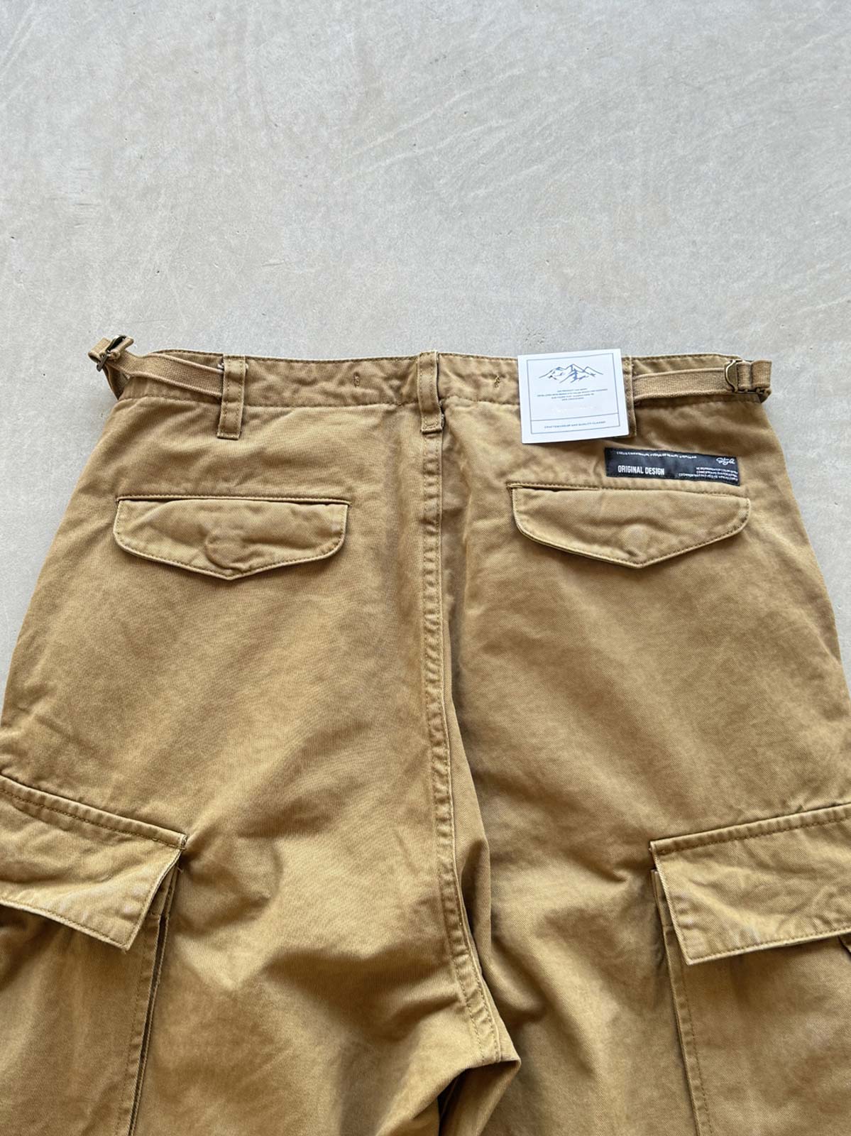 製品画像: 重厚なウォッシュ加工コットン100%を使用した多ポケットワークパンツ全体の外観、カーキ、グレー、ブラックの3色展開。
詳細画像: 純銅製YKKジッパーや調節可能なウエストバックル、裾のドローストリングディテールのクローズアップ。