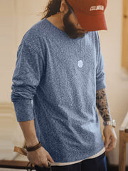 詳細画像: カチオン糸による独特な風合いとヴィンテージ感漂うブラウンのレザーパッチのディテールをクローズアップした画像。見た目の個性と高級感を強調。
モデル画像: 両面フリース起毛 長袖Tシャツを着用したモデルが、リラックスしたカジュアルコーディネートを披露。インナーとしても単独でも活躍するアイテム。