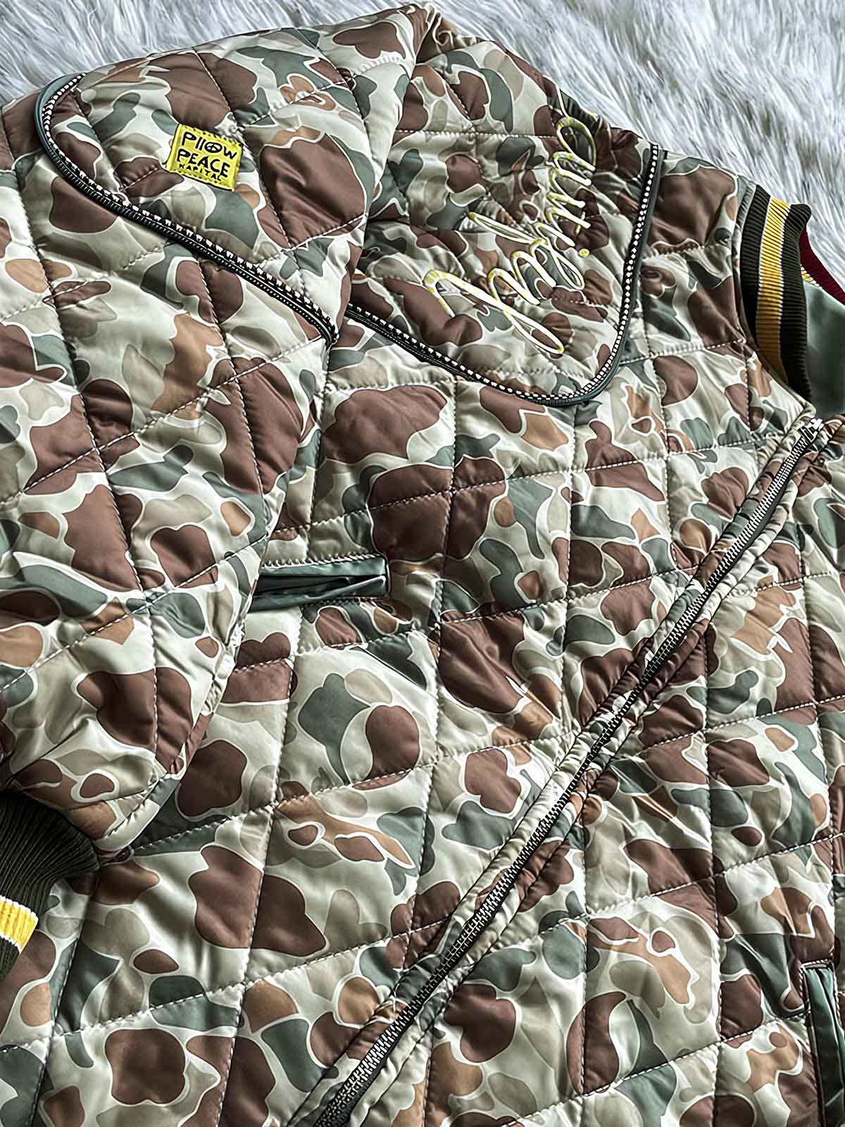 製品画像
「カモフラージュ刺繍ベースボールジャケット全体像。迷彩柄と平和を象徴する刺繍デザインが特徴のレトロスタイルジャケット」
詳細画像
「カモフラージュ刺繍ベースボールジャケットの袖部分の刺繍ディテールとストライプリブ仕様ポケットのクローズアップ。デザイン性と実用性を両立」