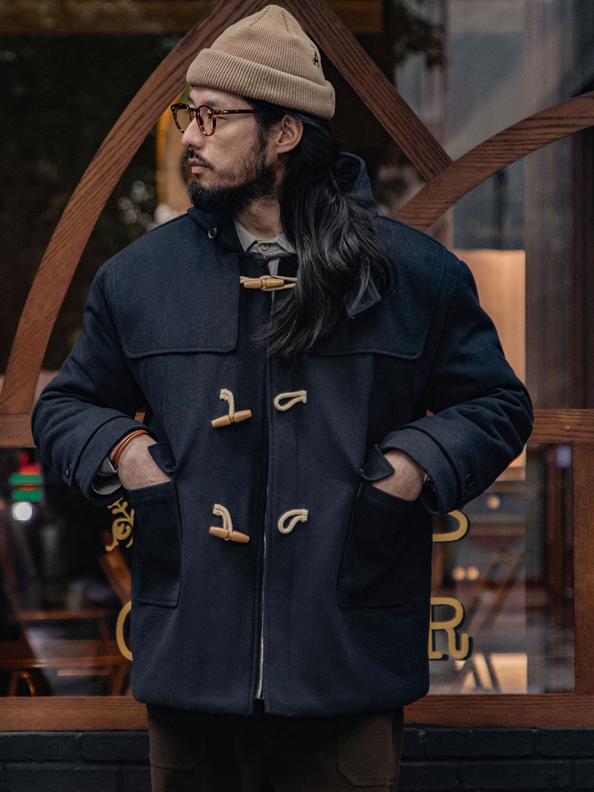 詳細画像
WWI時代のデザインを反映した木製トグルボタンと取り外し可能なストームフラップのディテール。160gウォッシャブルコットン中綿が暖かさと快適さを提供。
モデル画像
モデルがウール混ロング丈フード付きダッフルコートを着用し、冬のヴィンテージ通勤スタイルを演出。クラシックなデザインと現代的な機能性を融合した冬の必須アイテム。