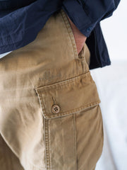 詳細画像
バティック染めによる独特なテクスチャーや、取り外し可能なナイロンベルト、UTX Duraflexバックル、そして膝部分の補強パッチとYKK製カスタムジッパーのディテールショット。高い機能性が際立つ細部。
モデル画像
ヴィンテージミリタリーテイストの多ポケットロングパンツを着用したモデルが、アウトドアコーディネートを披露。裾ドローコードで調節したリラックスシルエットが魅力のスタイル。