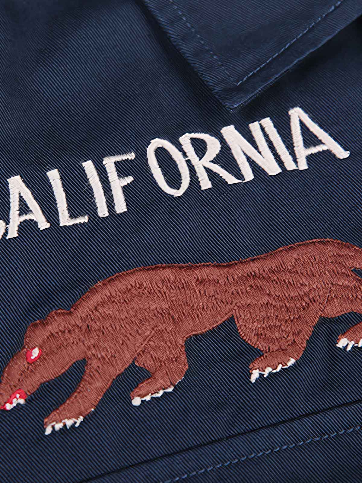 製品画像
厚手キャンバス生地で作られた刺繍キャンバスシャツの全体画像。1960年代のミリタリーユニフォームを再現したヴィンテージ感あるデザインが特徴。
詳細画像
フランス・ペリグーとアメリカ・カリフォルニア州をモチーフにした繊細な刺繍ディテールのクローズアップ。実用的なフラップ付きポケットも強調。