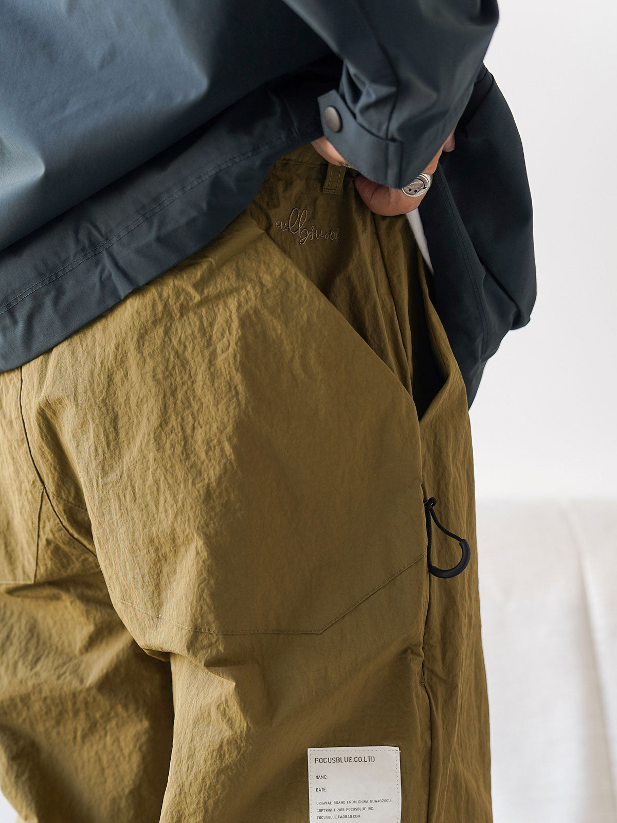 詳細画像
膝部分のダブルプリーツディテール、隠しポケット、UTX Duraflexドローコードエンド、そして反戦をテーマにしたデュポン紙ラベルのクローズアップ画像。機能性とディテールへのこだわりを強調。
モデル画像
シワ感テクスチャー素材のパラシュートパンツを着用したモデルが、機能性ジャケットと合わせたミリタリースタイルを披露。ワイドシルエットが快適さとトレンド感を演出。