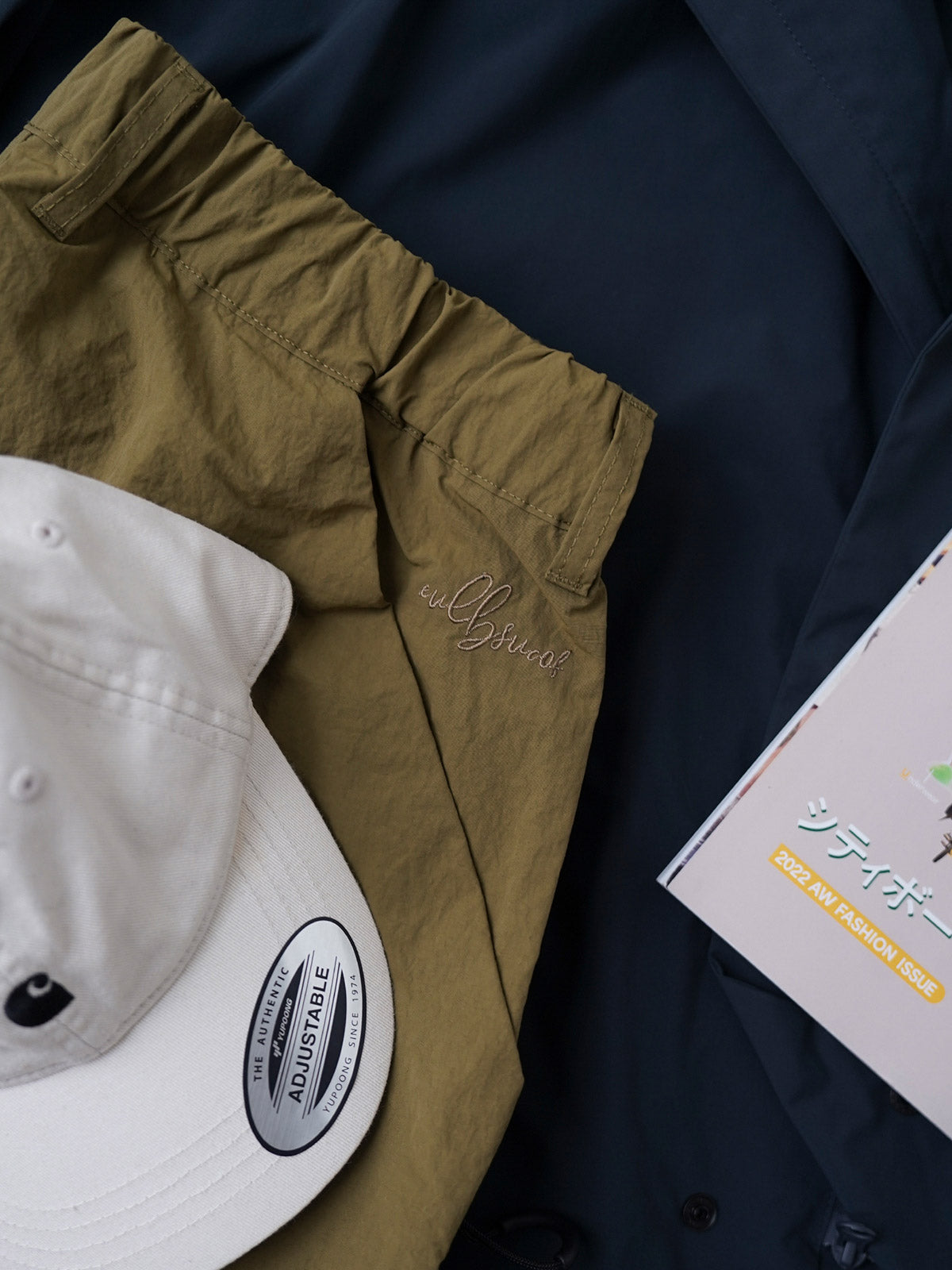 製品画像
撥水性に優れたナイロン素材を使用し、シワ感テクスチャーが特徴のミリタリーパラシュートパンツ全体画像。ダブルプリーツと多機能ポケットを備えたクラシックなシルエットが際立つデザイン。
詳細画像
膝部分のダブルプリーツディテール、隠しポケット、UTX Duraflexドローコードエンド、そして反戦をテーマにしたデュポン紙ラベルのクローズアップ画像。機能性とディテールへのこだわりを強調。