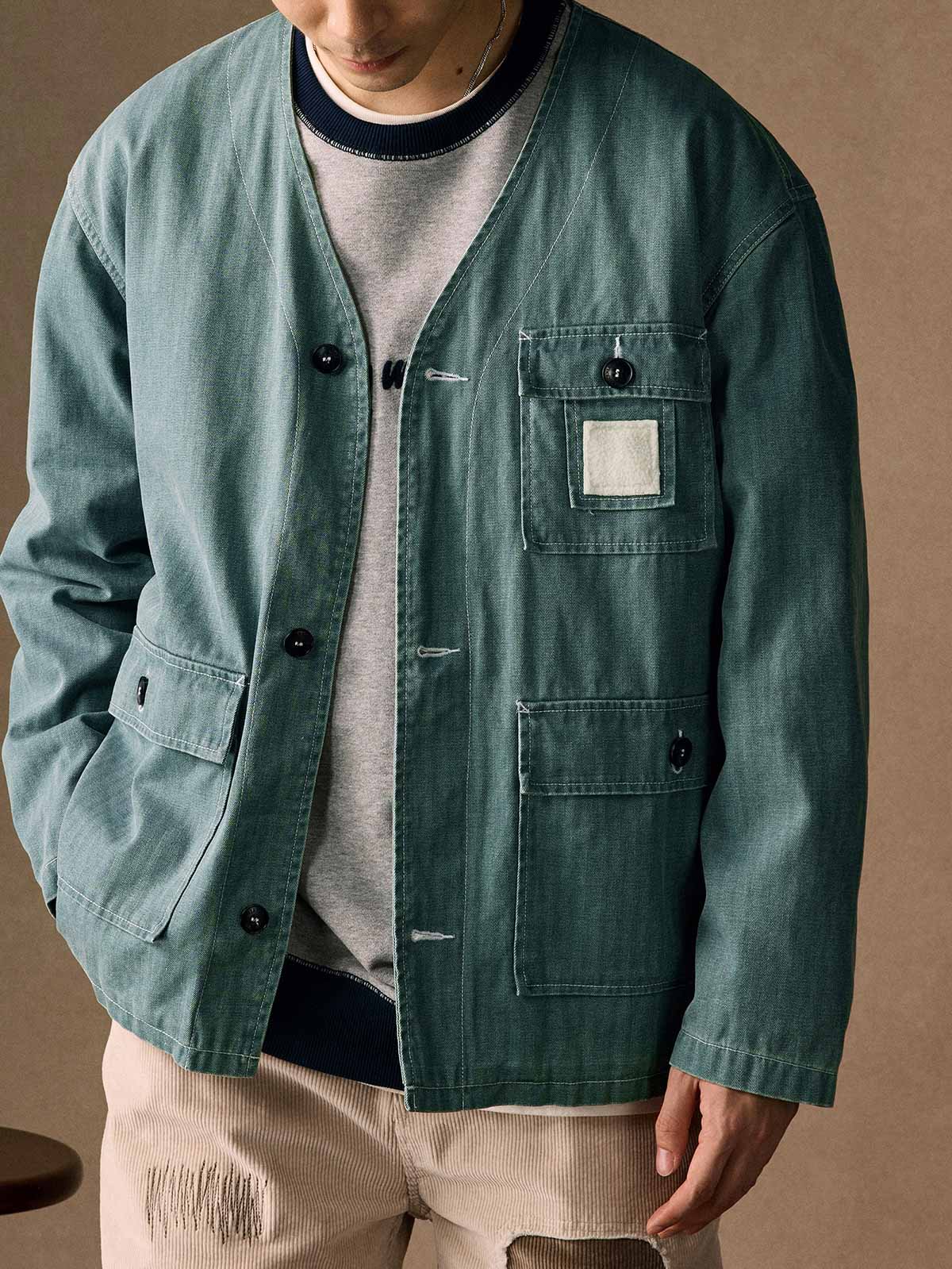 詳細画像
「ヘリンボーン生地のしっかりとしたテクスチャーとウォッシュ加工によるユーズド感、胸ポケットのラムウール織りディテールをクローズアップ。」
モデル画像
「インナーにシャツを合わせて着用したノーカラーフィッシングジャケット。ルーズフィットのシルエットがレイヤードスタイルを引き立てるヴィンテージ風コーディネート。」