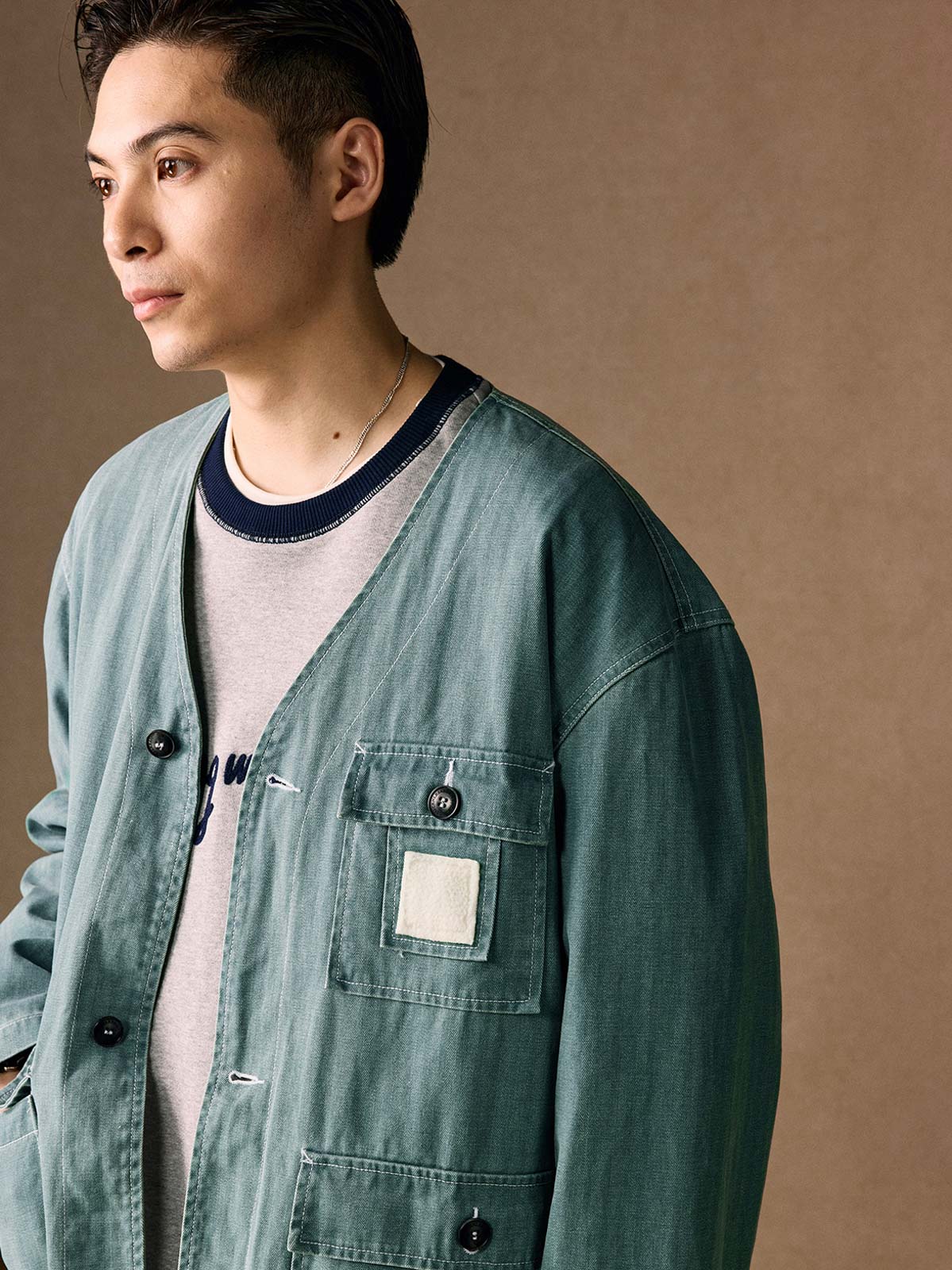 詳細画像
「ヘリンボーン生地のしっかりとしたテクスチャーとウォッシュ加工によるユーズド感、胸ポケットのラムウール織りディテールをクローズアップ。」
モデル画像
「インナーにシャツを合わせて着用したノーカラーフィッシングジャケット。ルーズフィットのシルエットがレイヤードスタイルを引き立てるヴィンテージ風コーディネート。」