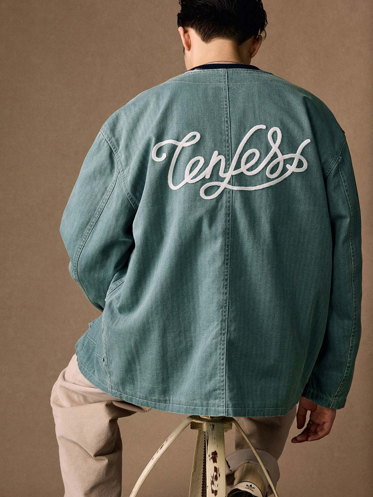 詳細画像
「ヘリンボーン生地のしっかりとしたテクスチャーとウォッシュ加工によるユーズド感、胸ポケットのラムウール織りディテールをクローズアップ。」
モデル画像
「インナーにシャツを合わせて着用したノーカラーフィッシングジャケット。ルーズフィットのシルエットがレイヤードスタイルを引き立てるヴィンテージ風コーディネート。」