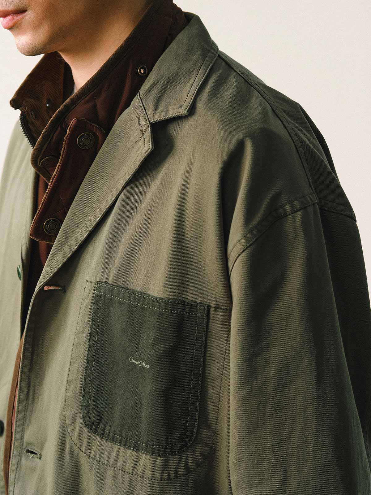 詳細画像
「バイカラーの胸ポケットやアシンメトリーな複合ポケットのディテールクローズアップ。酵素ウォッシュ加工による自然な色褪せ感と軽い起毛感が特徴。」
モデル画像
「モデルがアーミーグリーンのウォッシュ加工ワークテイストテーラードジャケットをデニムと合わせたヴィンテージ風カジュアルワークコーディネート。」