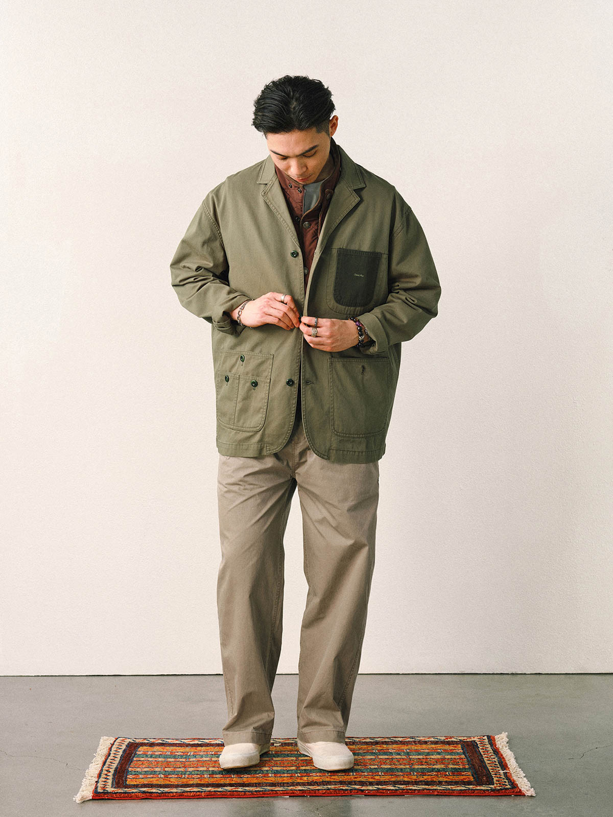 詳細画像
「バイカラーの胸ポケットやアシンメトリーな複合ポケットのディテールクローズアップ。酵素ウォッシュ加工による自然な色褪せ感と軽い起毛感が特徴。」
モデル画像
「モデルがアーミーグリーンのウォッシュ加工ワークテイストテーラードジャケットをデニムと合わせたヴィンテージ風カジュアルワークコーディネート。」