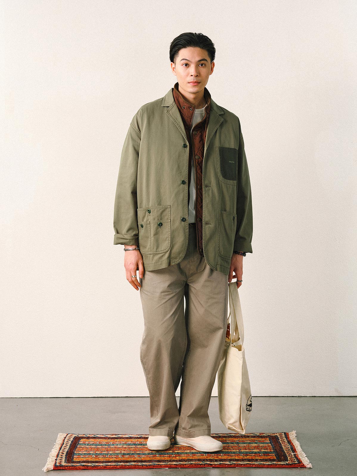 詳細画像
「バイカラーの胸ポケットやアシンメトリーな複合ポケットのディテールクローズアップ。酵素ウォッシュ加工による自然な色褪せ感と軽い起毛感が特徴。」
モデル画像
「モデルがアーミーグリーンのウォッシュ加工ワークテイストテーラードジャケットをデニムと合わせたヴィンテージ風カジュアルワークコーディネート。」