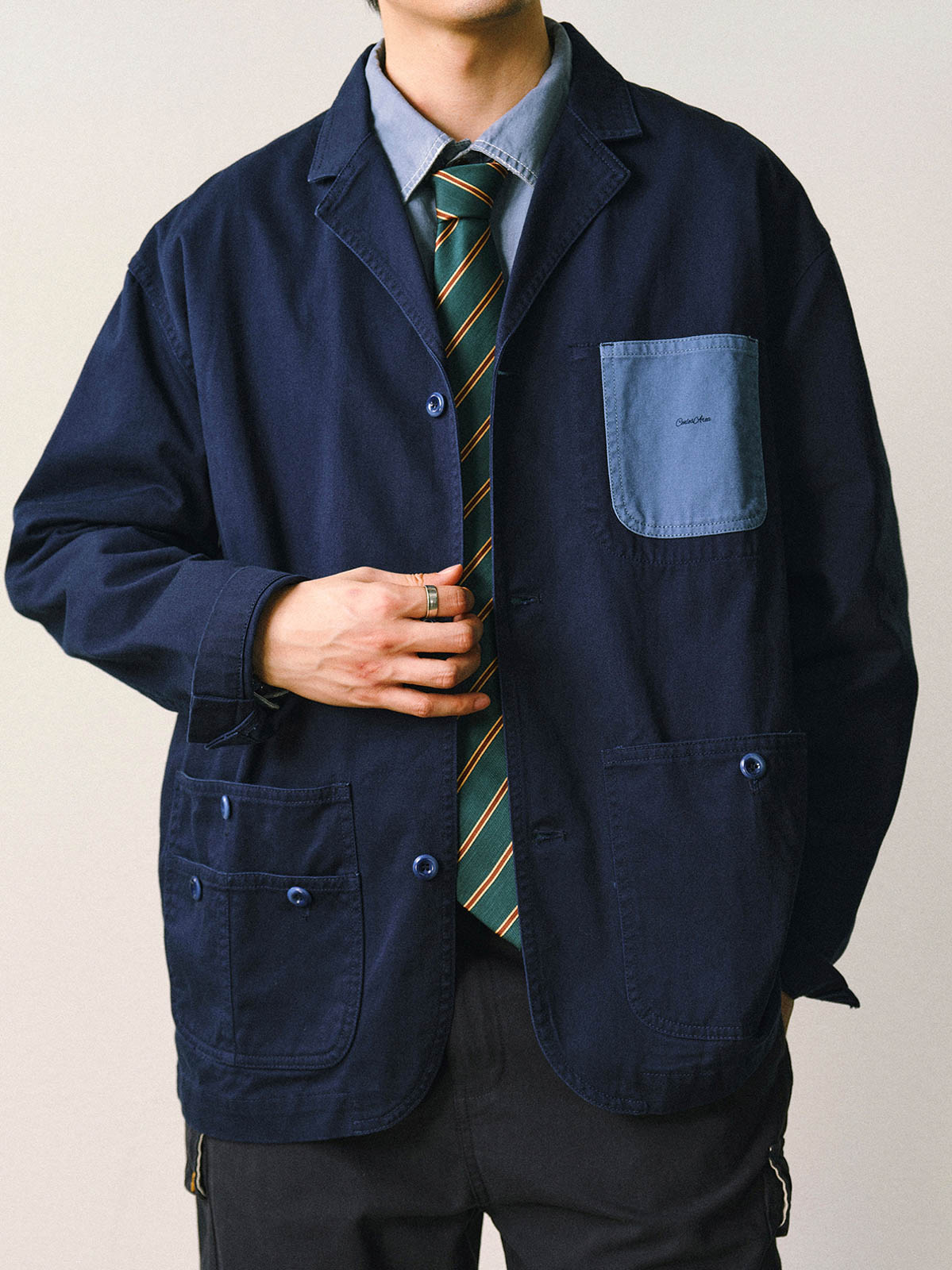 詳細画像
「バイカラーの胸ポケットやアシンメトリーな複合ポケットのディテールクローズアップ。酵素ウォッシュ加工による自然な色褪せ感と軽い起毛感が特徴。」
モデル画像
「モデルがアーミーグリーンのウォッシュ加工ワークテイストテーラードジャケットをデニムと合わせたヴィンテージ風カジュアルワークコーディネート。」