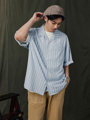 モデルが教会建築ストライプのハワイアンシャツを着用し、フラットフロントパンツとカジュアルシューズでスタイリング。リラックスしたビーチリゾートスタイルを演出。