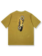 デザートイエローの背景にサボテンと野生の花の刺繍が施された「デザートフラワー」半袖Tシャツの正面ビュー。ゆったりとしたフィット感で、夏の日常着に理想的です。