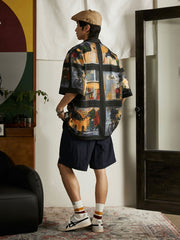 モデルが雨の日の街景油画風ハワイアンシャツを着用し、ジーンズとキャンバスシューズでコーディネート。ダークトーンのプリントがカジュアルながらもアートフルなスタイルを提供し、週末のリラックスしたアウトドアやカジュアルな街歩きに適しています。