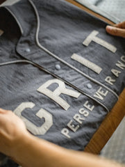 メテオライトグレーのベースボールシャツ、胸元に「GRIT PERSEVERANCE」フェルトパッチが施されている