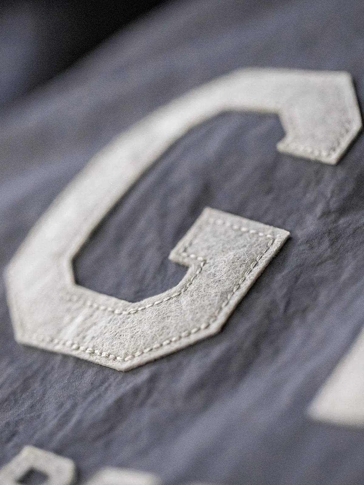 メテオライトグレーのベースボールシャツ、胸元に「GRIT PERSEVERANCE」フェルトパッチが施されている