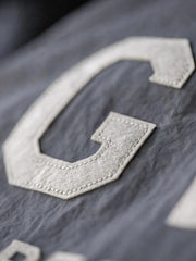 メテオライトグレーのベースボールシャツ、胸元に「GRIT PERSEVERANCE」フェルトパッチが施されている