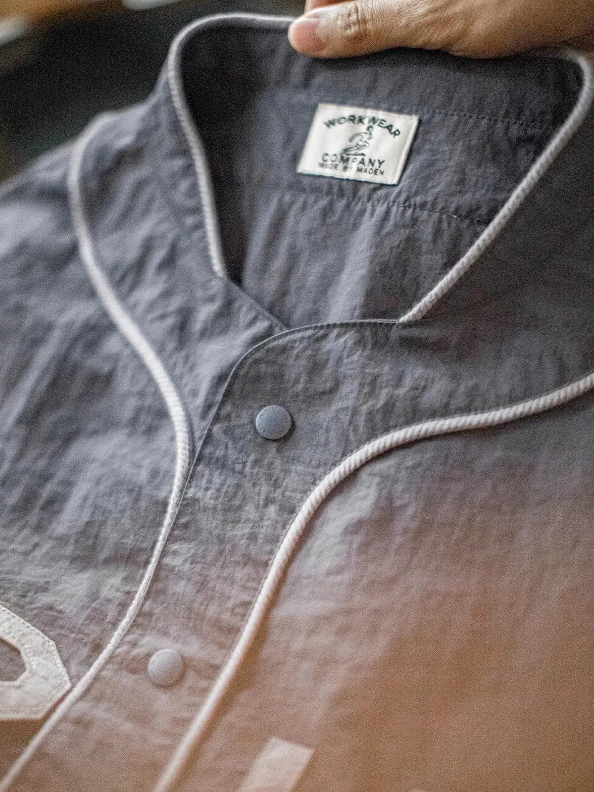 メテオライトグレーのベースボールシャツ、胸元に「GRIT PERSEVERANCE」フェルトパッチが施されている