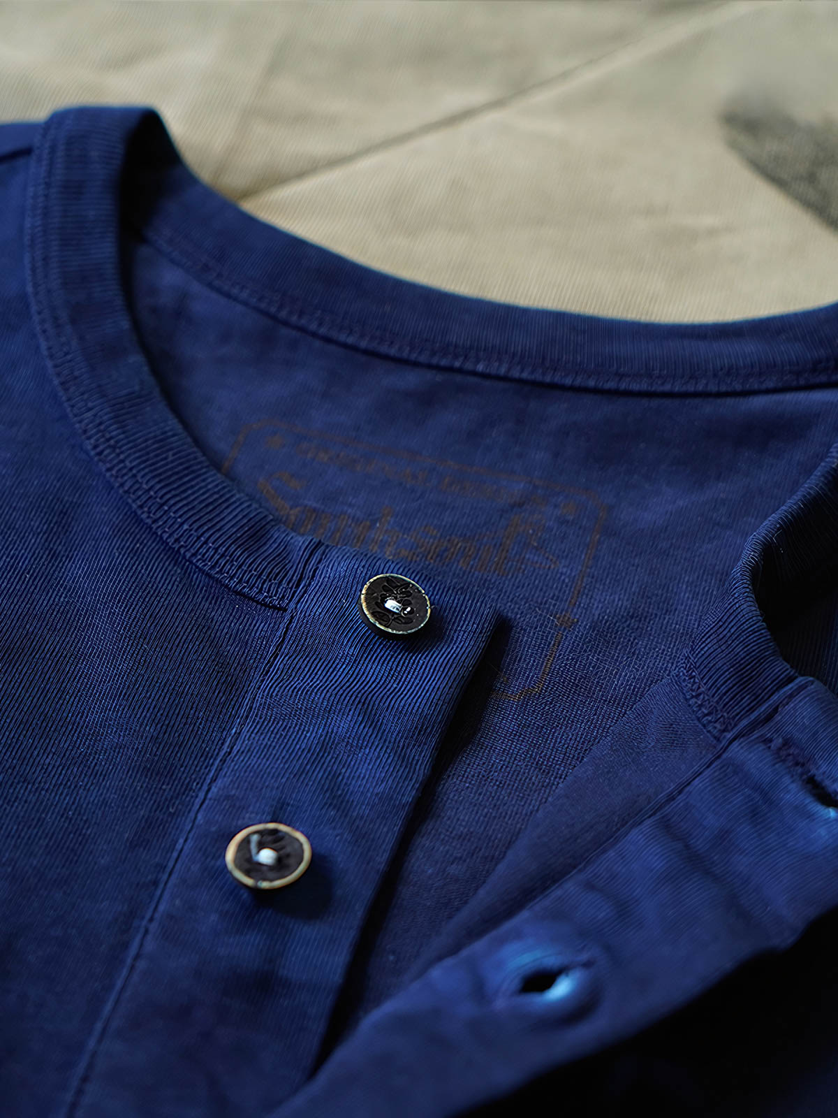 製品画像
伝統的な藍染め技法を施したヘンリーネック長袖Tシャツの全体画像。260gの厚手コットン素材とクラシックなデザインが際立つ一枚。
詳細画像
藍染めによる独特なグラデーションと、染色されたコットン縫い目の細部を強調したクローズアップ画像。1点ごとに異なる色合いや職人の手作り感が魅力。