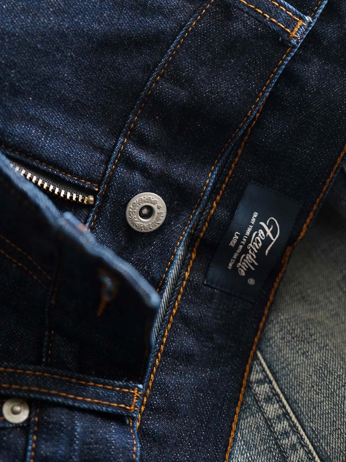 製品画像
「14OZ赤耳デニムパンツ全体イメージ。旧式織機で織られたクラシックな狭幅赤耳デニム生地とインディゴ染めの高品質な仕上がりが特徴。」
詳細画像
「赤耳セルビッジ、インディゴ染め鹿革パッチ、月桂冠モチーフのドーナツボタン、カスタムYKKジッパーなど、クラシックで高級感のあるディテールのクローズアップ。」