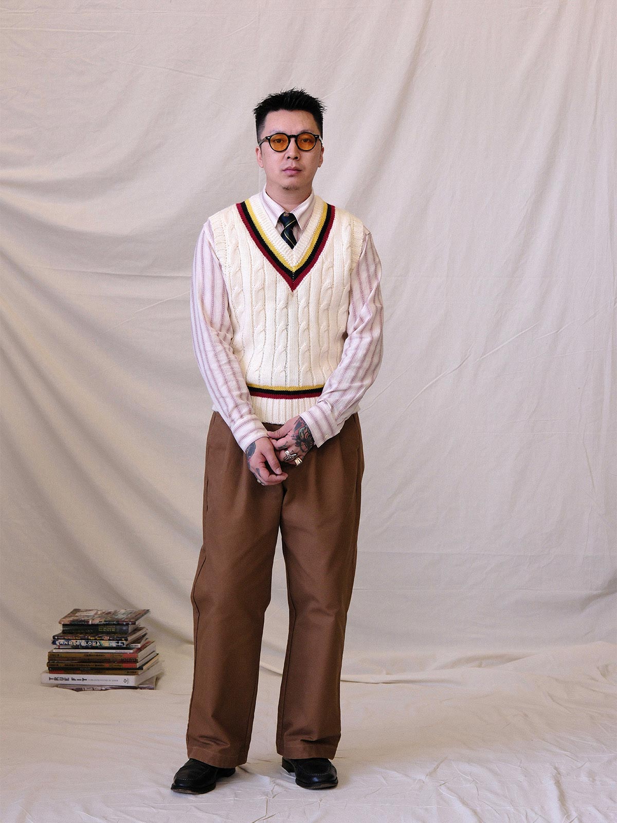 詳細画像:
ツイスト編みの編み目とカラーコンビネーションストライプのディテール。Vネックとストライプデザインが加える視覚的なレイヤード感を強調した画像。
モデル画像:
モデルがシャツと重ね着したVネック クリケットニットベストを着用し、英国風のアカデミックスタイルを演出している画像。