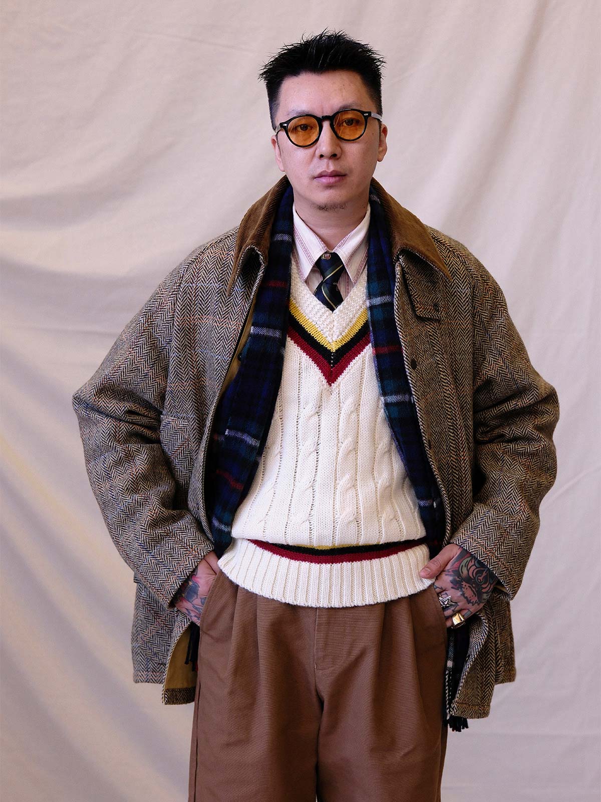 詳細画像:
ツイスト編みの編み目とカラーコンビネーションストライプのディテール。Vネックとストライプデザインが加える視覚的なレイヤード感を強調した画像。
モデル画像:
モデルがシャツと重ね着したVネック クリケットニットベストを着用し、英国風のアカデミックスタイルを演出している画像。