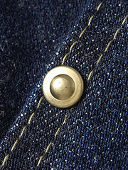 製品画像
1940年代戦時中のS101Jを忠実に再現したリバイバルデニムジャケットの正面デザイン。14オンスのセルビッジデニムとヴィンテージ感溢れるインディゴブルーの色合いが特徴
詳細画像
鉄製の月桂(ゲッケイ)ボタンと真鍮製フライングソーサーリベットのディテール。経年変化による自然な錆びが美しく、クラシックなワークウェアの魅力を引き立てる精密な縫製