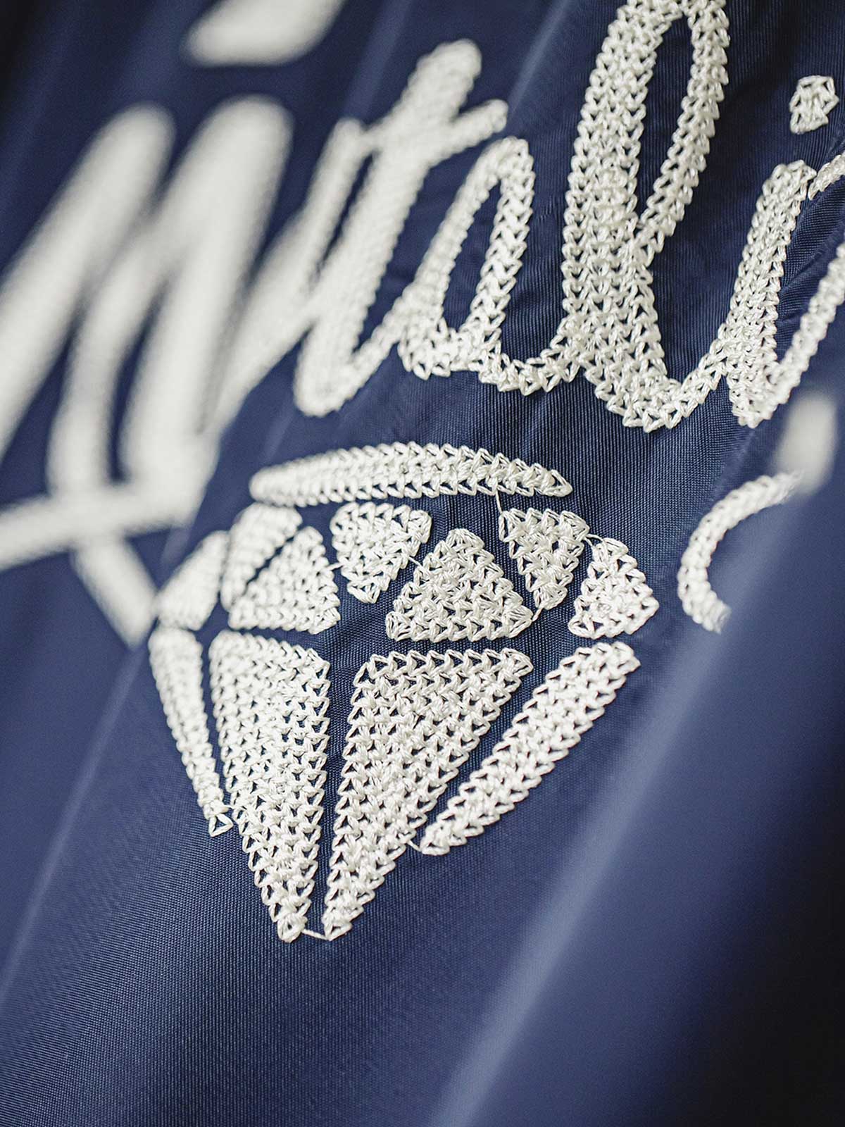 製品画像
「ミッドナイトネイビーのヴィンテージ感あふれる軽量刺繍ワークジャケット正面展示写真。150gナイロン素材とクラシックなフレーザータータンチェック裏地を組み合わせた秋冬の個性的な一着。」
詳細画像
「背面に施された『Hustle is Mentality』スローガンとダイヤモンド型チェーン刺繍のディテール写真。クラシックなデザインと挑戦者の精神を象徴するヴィンテージスタイルのジャケット。」