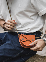 無地シャツとカジュアルパンツを組み合わせ、二重フラップ植物タンニン鞣しレザーのサドルバッグを肩にかけたモデル。