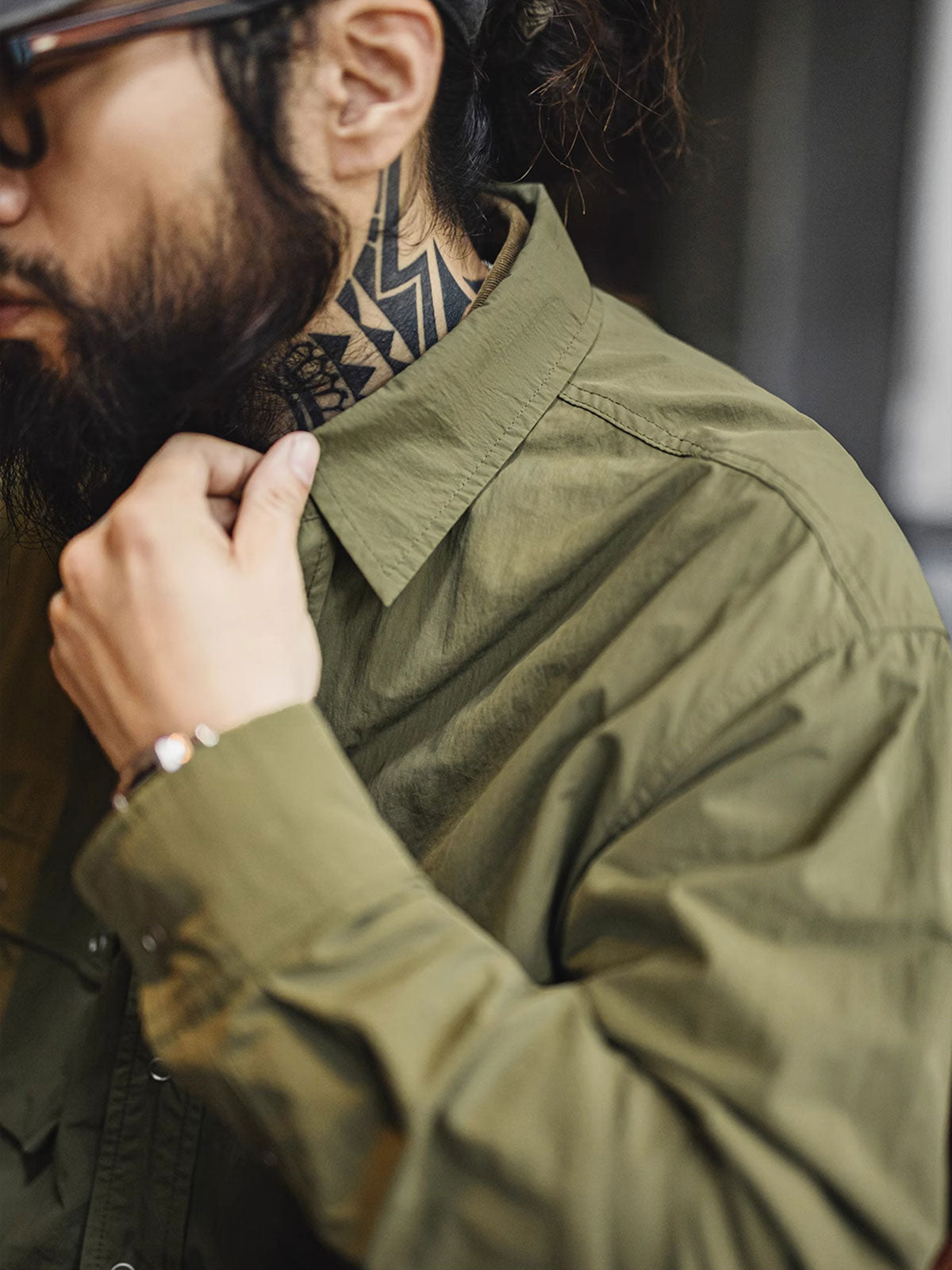 詳細画像
マットな5本爪スナップボタンと、戦地カメラマンの実用性を考慮して配置された3つのフラップ付きポケットのディテール特写。独自のシワ加工が施され、経年変化を楽しめる仕上がり
モデル画像
ノマドジャーナリストシャツを着用したコーディネート例。肩幅にゆとりを持たせたルーズフィットデザインが、ミリタリースタイルと現代的なワークウェアとしての快適さを融合したスタイリングを演出