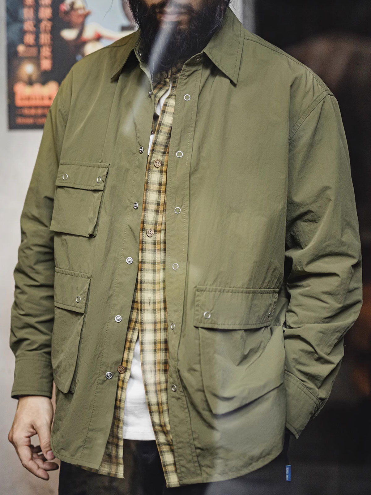 詳細画像
マットな5本爪スナップボタンと、戦地カメラマンの実用性を考慮して配置された3つのフラップ付きポケットのディテール特写。独自のシワ加工が施され、経年変化を楽しめる仕上がり
モデル画像
ノマドジャーナリストシャツを着用したコーディネート例。肩幅にゆとりを持たせたルーズフィットデザインが、ミリタリースタイルと現代的なワークウェアとしての快適さを融合したスタイリングを演出