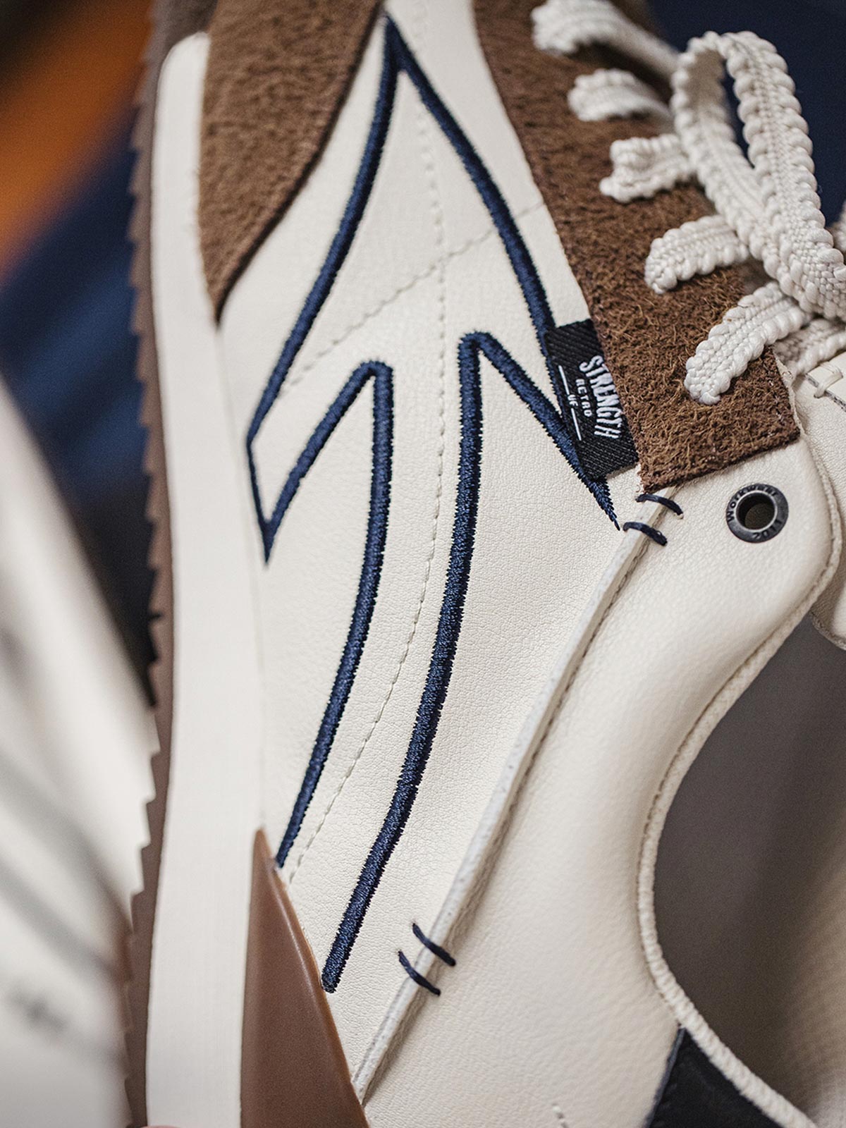 製品画像: 60Sピースバスケットシューズ正面展示画像。長毛感のあるスエード革と異素材のコンビネーションが特徴で、ネイビーブルーのエレクトリック刺繍矢印ロゴがサイドに配置されています。ヒール部分にはブラックの本革と金色のレトロな金属刻印が施され、独自のデザインを際立たせています。
詳細画像: 60Sピースバスケットシューズのアッパー部分、長毛感のあるスエード革と異素材のコンビネーションが美しく融合し、レトロ感を強調しています。金色のレトロ金属刻印と刺繍のディテールがシューズに高級感を与えています。