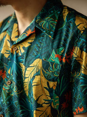 モデルがハワイアンプリント半袖シャツを着用し、無地のカジュアルパンツとシンプルなサンダルを合わせたスタイルでポーズ。明るく夏らしいカジュアルスタイルを演出。