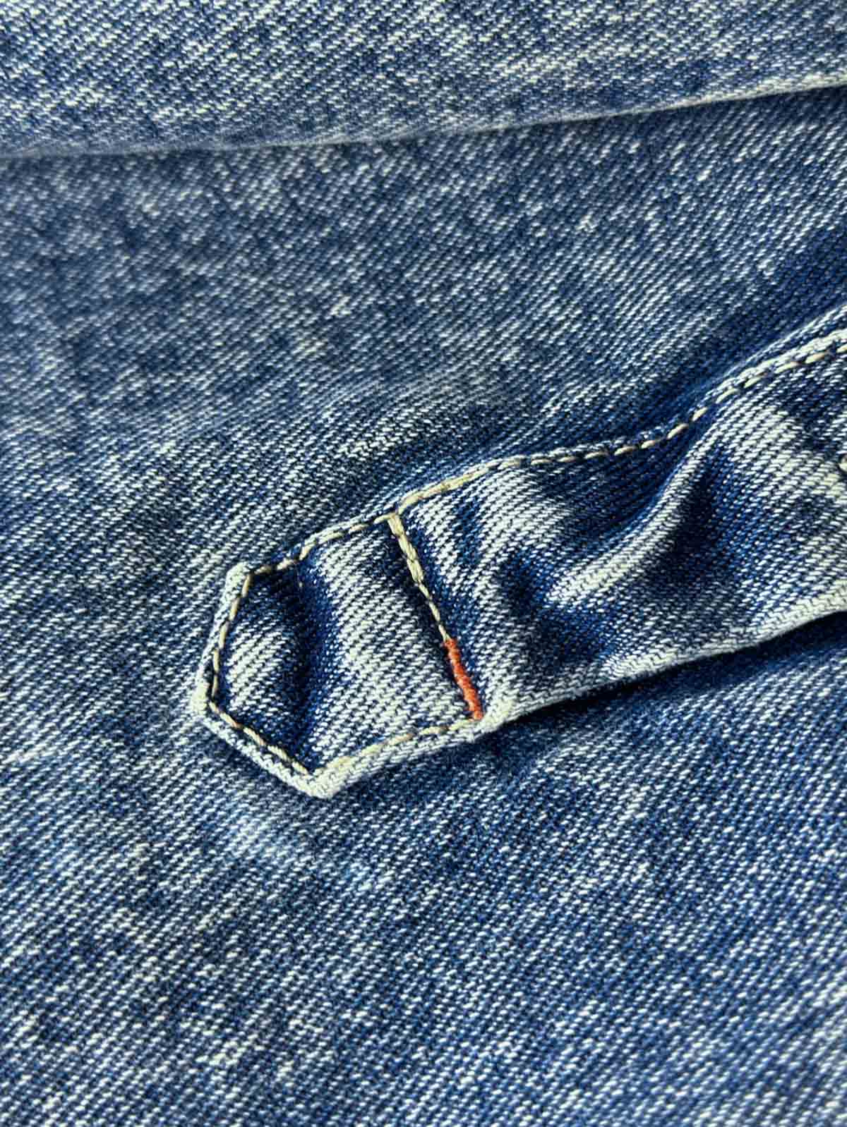 デニムシャツの袖口にあるタイスリーブノットとポケットのステッチラインのクローズアップ。手作業の細かなデテールが強調されている。