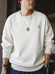 詳細画像: 胸元に配置された精巧な「ピースマーク」プリントとクラシックなリブ仕様のネックや裾をクローズアップした画像。保温性と耐久性を兼ね備えた裏起毛素材が際立つ。
モデル画像: 裏起毛クルーネックスウェットを着用したモデルが、スニーカーとブラックパンツを合わせたカジュアルな秋冬スタイルを提案。快適さと個性を両立したコーディネートが魅力的。