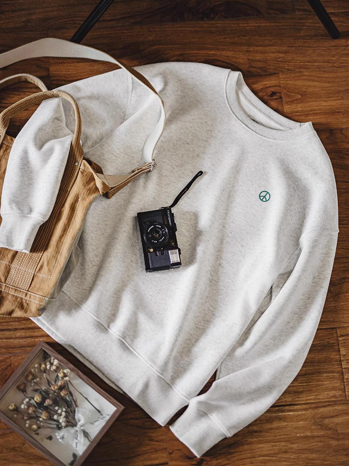 製品画像: ライトグレーの裏起毛クルーネックスウェット全体イメージ画像。リラックスシルエットとドロップショルダーデザインが特徴で、胸元には「ピースマーク」プリントがアクセント。単独でもインナーとしても活躍する秋冬の万能アイテム。
詳細画像: 胸元に配置された精巧な「ピースマーク」プリントとクラシックなリブ仕様のネックや裾をクローズアップした画像。保温性と耐久性を兼ね備えた裏起毛素材が際立つ。