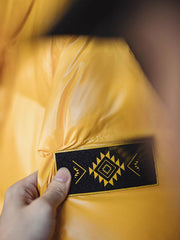 製品画像
鮮やかなイエローのハイカラー・ダウンジャケット、80年代のパニナーロスタイルを再現、撥水性4級のナイロン防水素材、クラシックなデザインと高襟が特徴のジャケット
詳細画像
六角形ジッパーチャームとインディアン柄刺繍ディテール、鮮やかなイエローが際立つデザイン、防水加工されたナイロン素材とレトロなディテールのクローズアップ