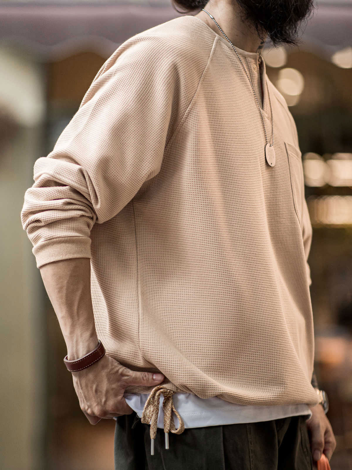 詳細画像
幾何学模様のワッフル編みテクスチャー、袖口のリブディテール、裾のドローストリングデザインを強調したクローズアップ画像。ラグランスリーブと切り替えデザインが動きと立体感を演出。
モデル画像
ヴィンテージ感漂うヘンリーネックシャツを着用したモデルが、ラグランスリーブによるリラックスシルエットを活かしたカジュアルスタイルを披露。快適な着心地と立体的な編み地が目を引くコーディネート。