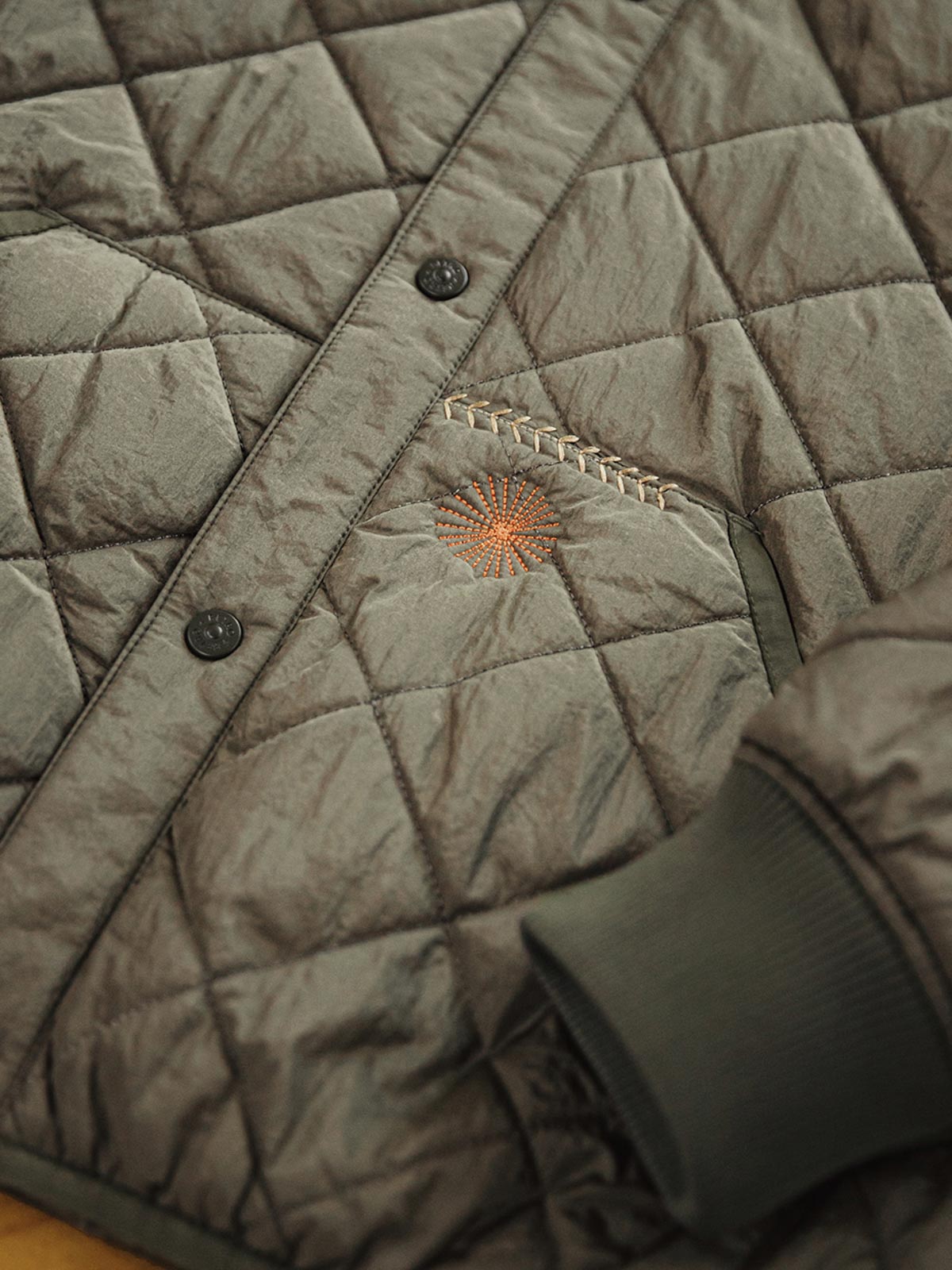 製品画像: レトロなキルティングベースボールジャケットデザイン中綿ジャケット正面展示画像。クラシックなベースボールジャケットのシルエットに、精緻なキルティング技術とオレンジ色の手刺繍ディテールが特徴。光沢ナイロン素材とポリエステル繊維で暖かさとスタイルを提供します。
詳細画像: キルティングベースボールジャケットのステッチポケットとオレンジ色の手刺繍風ディテールのクローズアップ。銅製スナップボタンのディテールと、均等に分布した中綿が暖かさを保証。