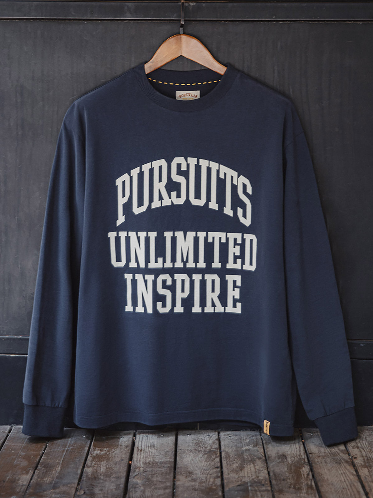 製品画像
「フロック加工ロゴプリントを大胆に施したアメリカンストリート風長袖Tシャツの全体画像。起毛加工の柔らかなコットン素材が快適な着心地を提供。」
詳細画像
「フロック加工のロゴプリントと起毛素材の滑らかな肌触りをクローズアップ。背面の刺繍アーチロゴや裾のラバープリントタグもディテールとして特徴的。」