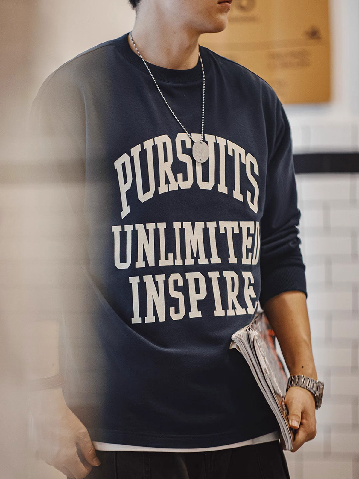 詳細画像
「フロック加工のロゴプリントと起毛素材の滑らかな肌触りをクローズアップ。背面の刺繍アーチロゴや裾のラバープリントタグもディテールとして特徴的。」
モデル画像
「モデルがフロック加工ロゴプリント長袖Tシャツを着用し、ドロップショルダーとクルーネックが特徴のアメリカンストリートスタイルを披露。」