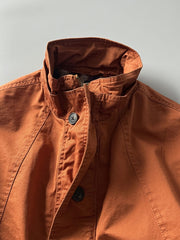 製品画像: オレンジカラーのダブルスタンドカラーワークジャケットの正面画像、実用的でスタイリッシュなデザインが特徴。
詳細画像: 袖部のジッパーポケットとスタンドカラーデザインのディテール画像、ワークスタイルを強調。