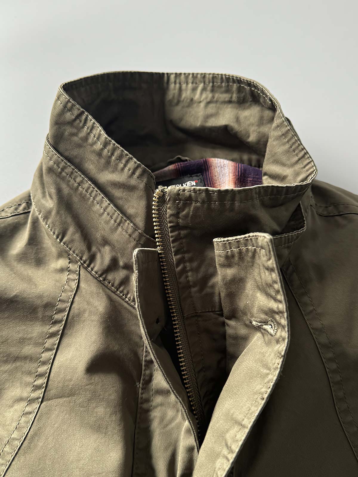 製品画像: オレンジカラーのダブルスタンドカラーワークジャケットの正面画像、実用的でスタイリッシュなデザインが特徴。
詳細画像: 袖部のジッパーポケットとスタンドカラーデザインのディテール画像、ワークスタイルを強調。