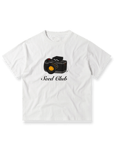コンリホワイトカメラプリントTシャツの全体像、クラシックなカメラモチーフとルーズフィットシルエットを示す。